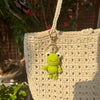 A handmade crochet frog keychain charm hanging from a beige knitted bag.