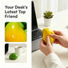 A hand holds a yellow mango-shaped squishy stress ball next to a laptop on a white desk.