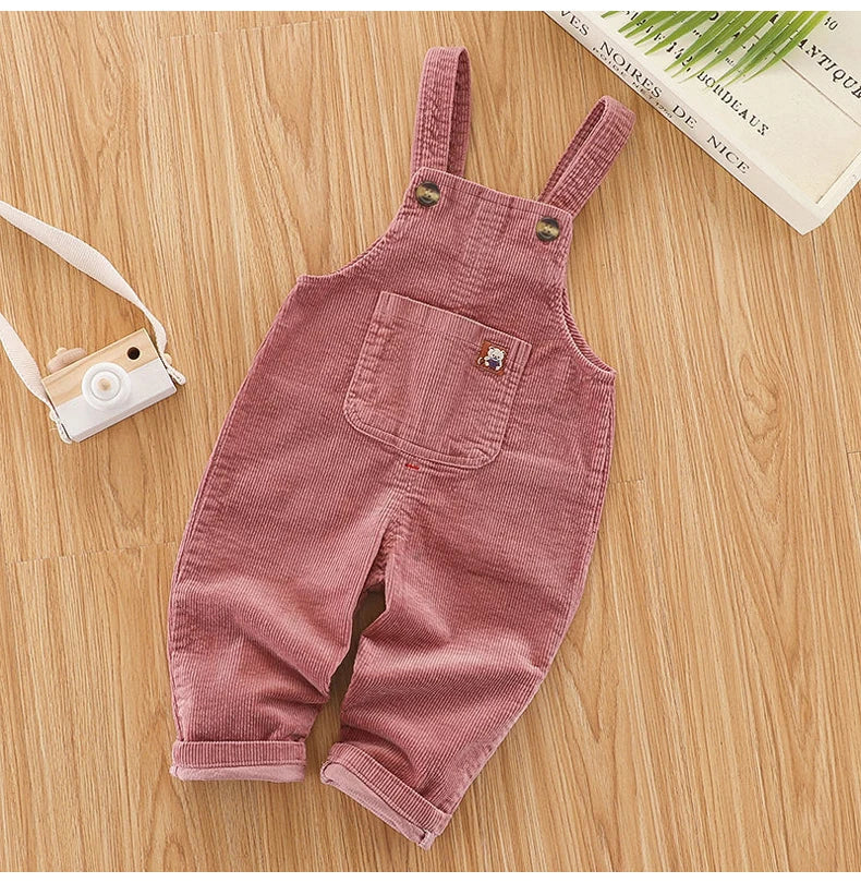 A pair of dusty rose corduroy baby cotton overalls with adjustable straps lying on a wooden surface.