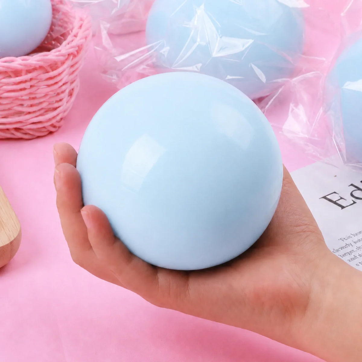 A hand holds a smooth, light blue squishy ball with clear plastic packaging on a pink background.