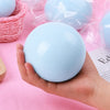 A hand holds a smooth, light blue squishy ball with clear plastic packaging on a pink background.