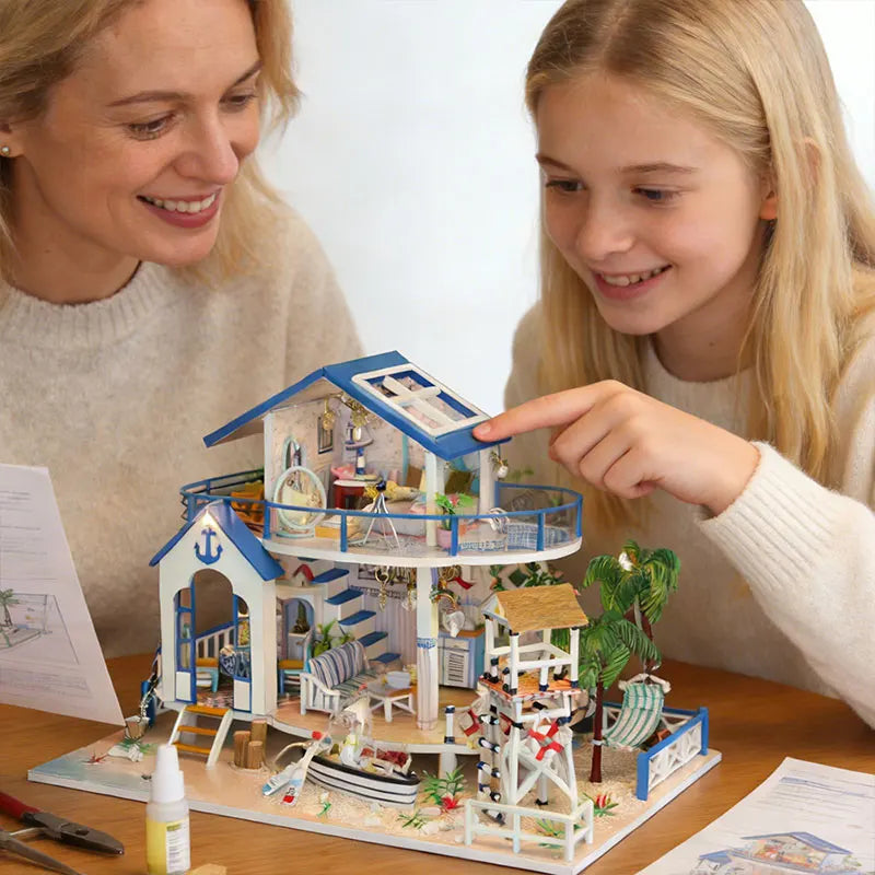 A woman and a girl look at a seaside beach house miniature dollhouse kit with an LED light.