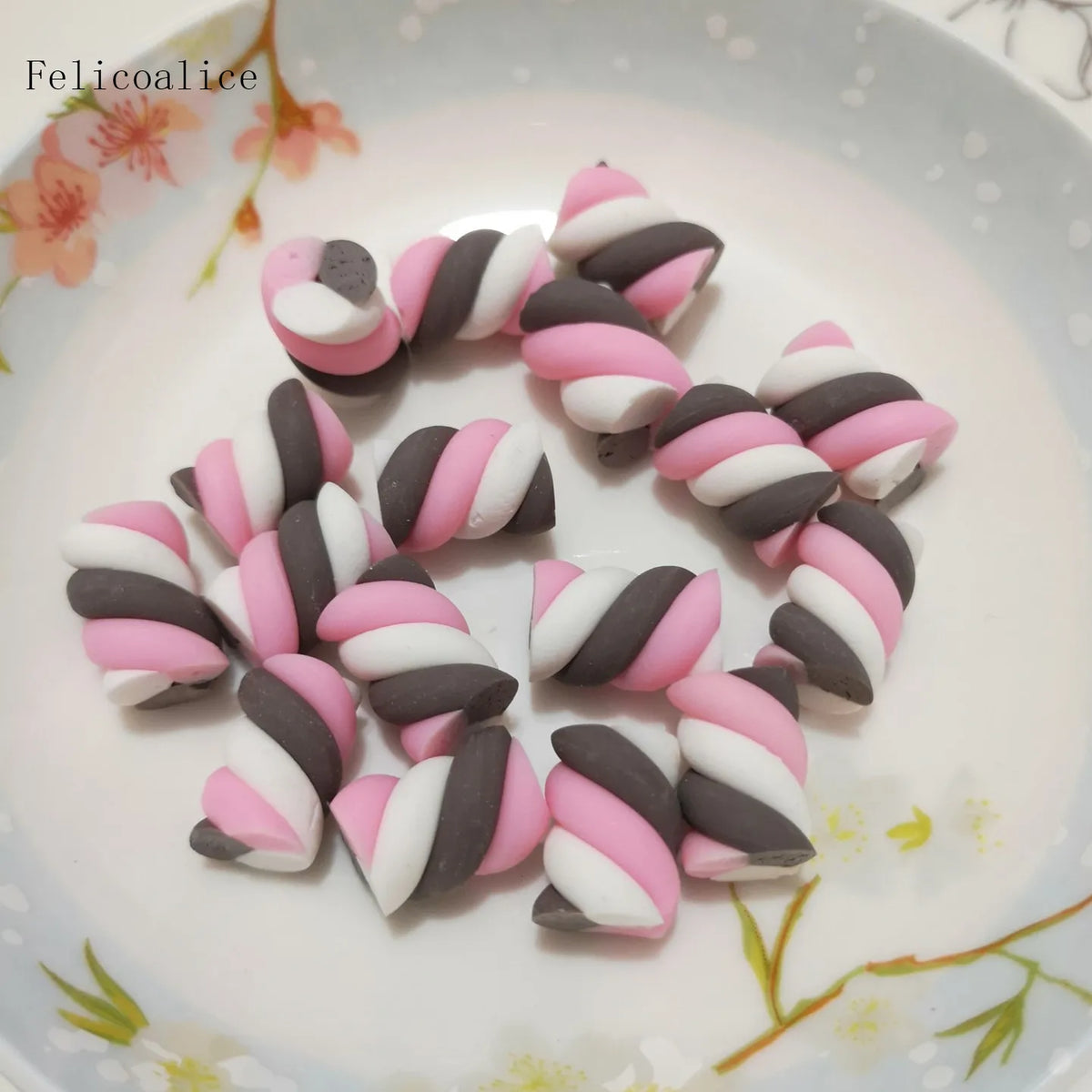 A collection of pink and white polymer clay marshmallow charms shaped like striped candies arranged on a white plate.