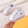 A pair of white canvas baby shoes with gray laces and a star detail are being held against a white knitted background.