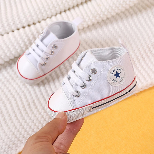 A pair of white canvas baby shoes with gray laces and a star detail are being held against a white knitted background.