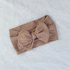 A beige knitted baby headband with a bow shown on a white textured fabric.