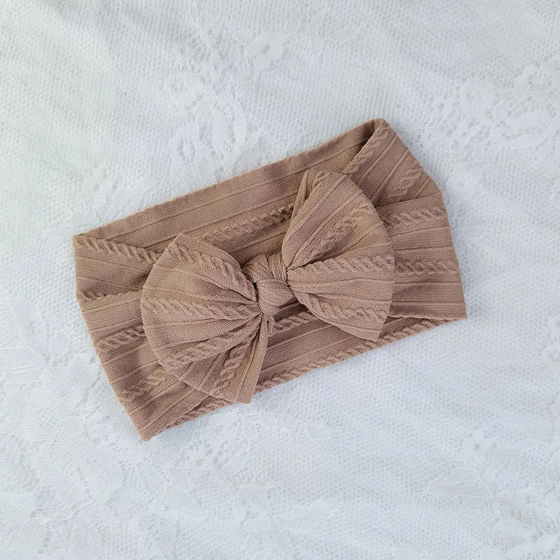 A beige knitted baby headband with a bow shown on a white textured fabric.