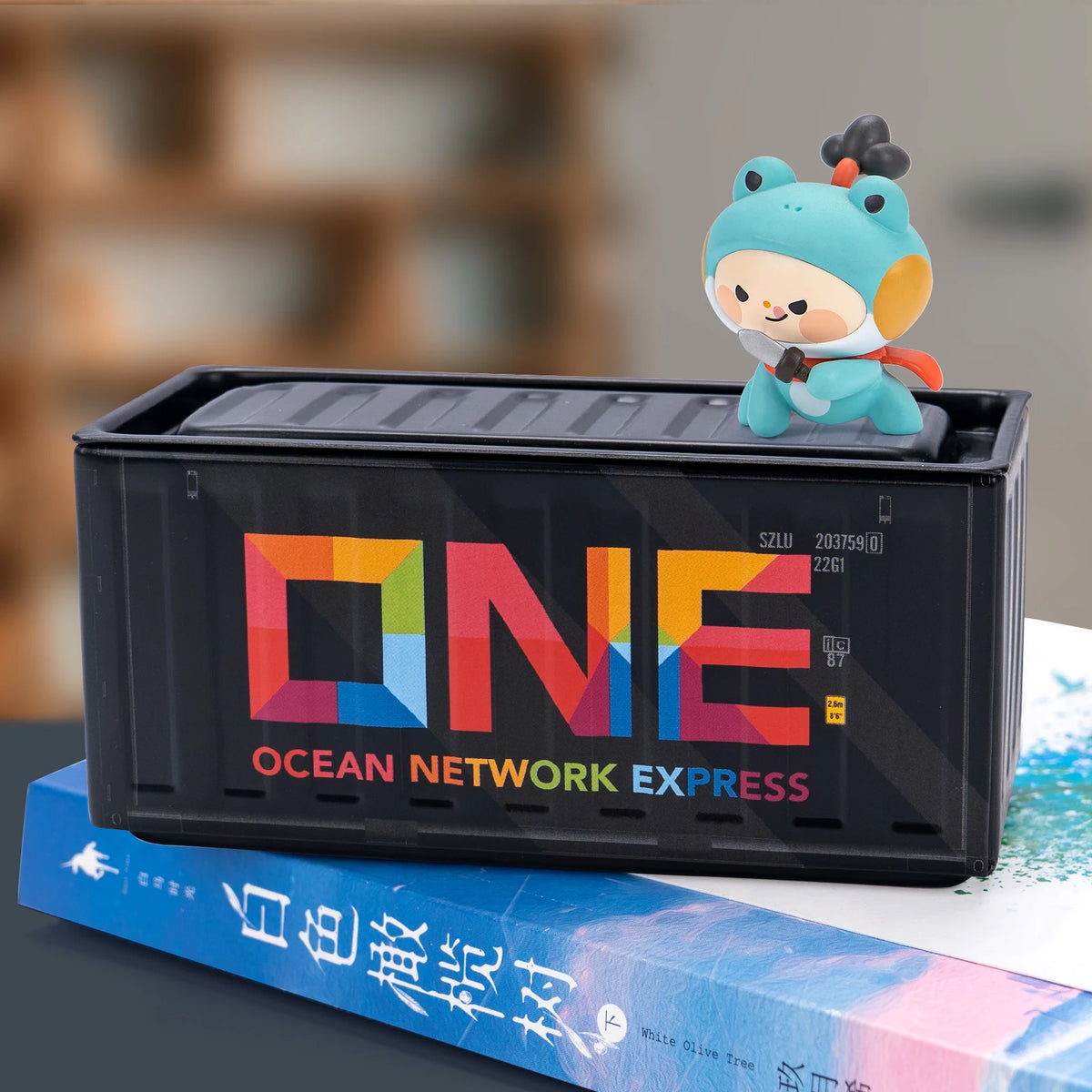 A small blue figurine sits on a black industrial iron storage box labeled Ocean Network Express next to a book.