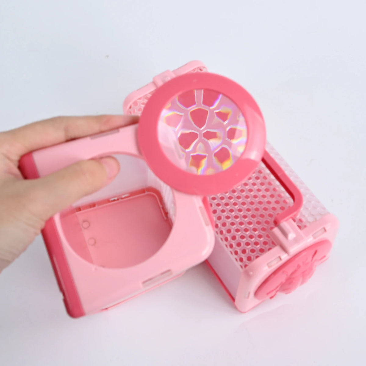 A hand holds a pink plastic insect catcher with a clear observation lens and a mesh cage.