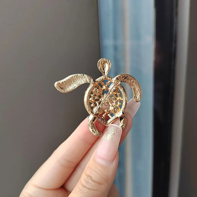 A close-up of a gold rhinestone turtle brooch pin being held by a hand against a light blue background.