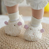 A pair of white, fluffy cartoon baby booties with pink ears and white socks resting on a woven mat.