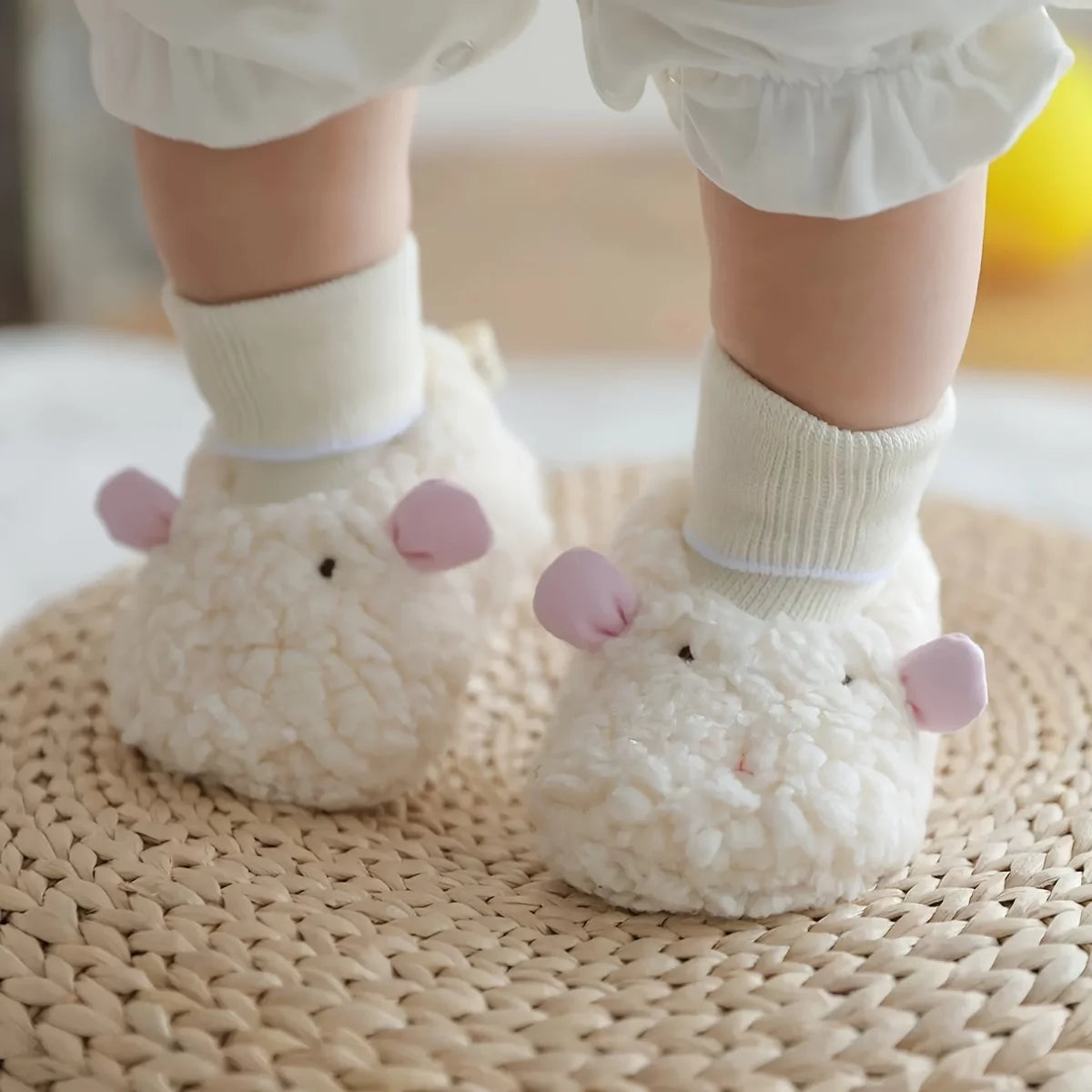 A pair of white, fluffy cartoon baby booties with pink ears and white socks resting on a woven mat.