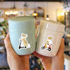 A hand holds two light green and off-white plastic containers featuring cartoon cats in wheelchairs.