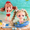 A pair of cute, pink-haired mini dolls with poseable joints sitting on a sandy surface near water.