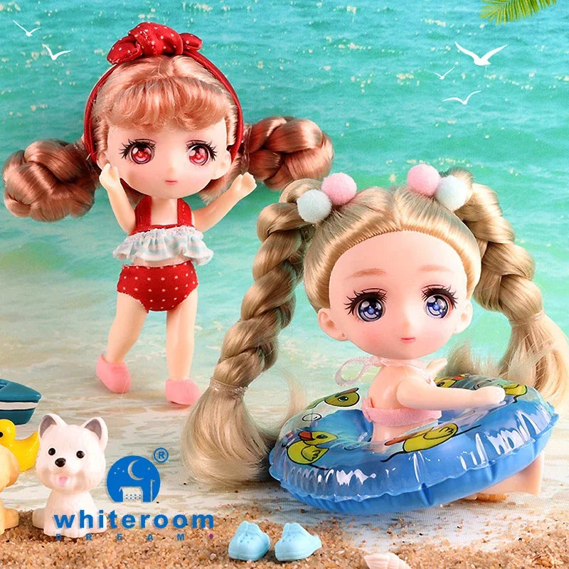 A pair of cute, pink-haired mini dolls with poseable joints sitting on a sandy surface near water.