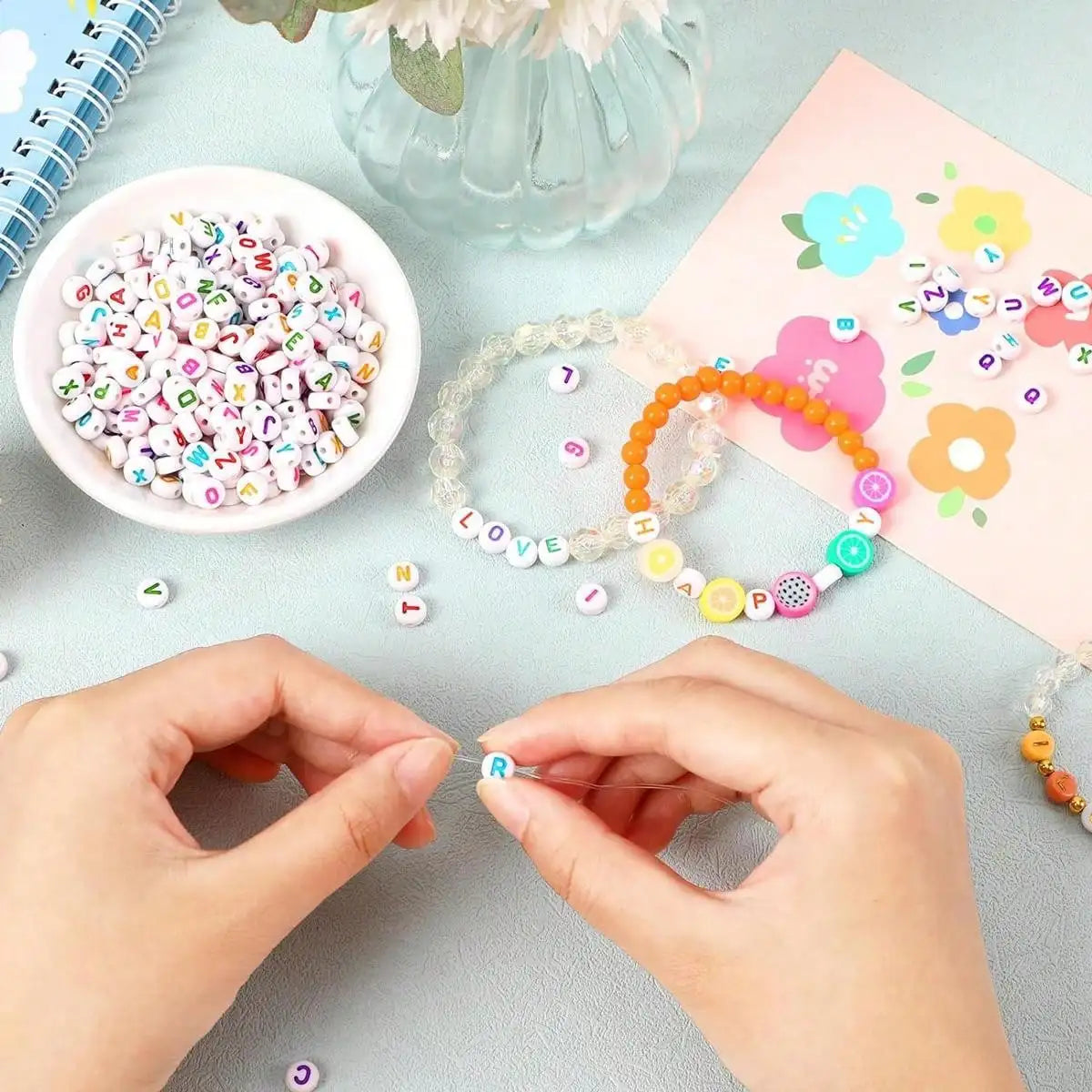 Hands are assembling letter and number acrylic beads for jewelry making on a light blue surface.