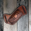 A brown leather knife sheath with a belt loop featuring a woven pattern rests on weathered wooden planks.