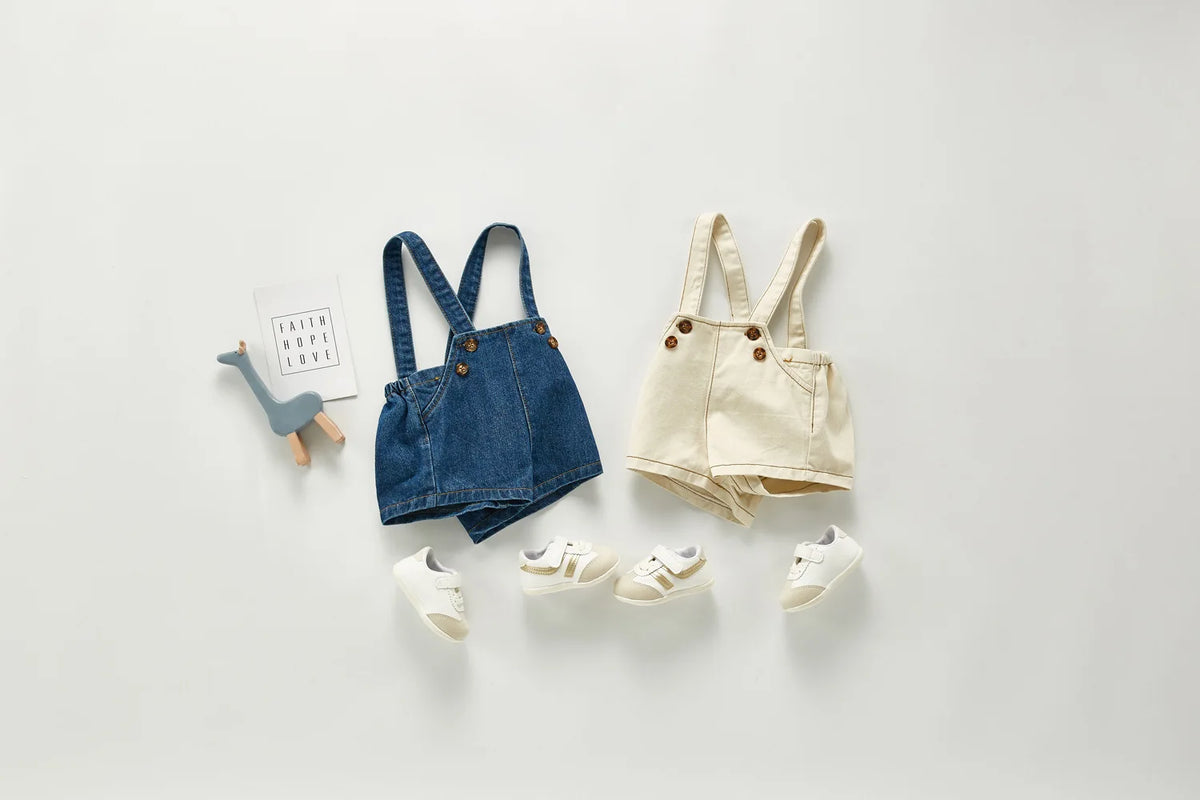 A denim romper for a toddler with pockets and matching light colored shoes displayed on a white background.