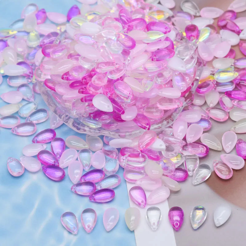 A pile of iridescent pink and purple water drop glass beads for jewelry making scattered on a light blue surface.