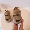 A hand holds a pair of beige toddler soft-soled sandals for boys and girls on a white surface.