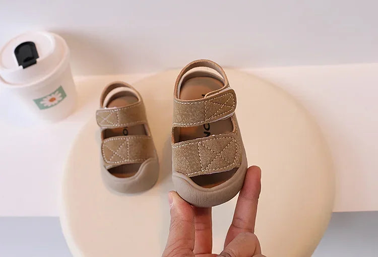 A hand holds a pair of beige toddler soft-soled sandals for boys and girls on a white surface.