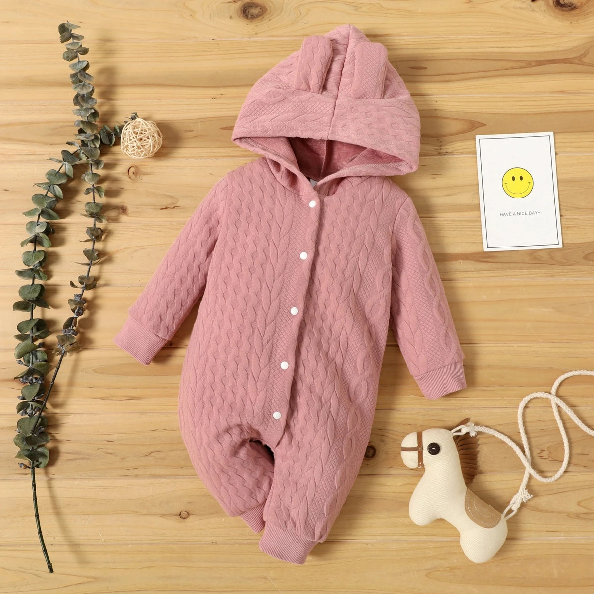 A pink quilted hooded baby romper lying on a wooden surface next to a eucalyptus branch and a smiling emoji card.