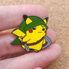 A yellow Pokémon enamel pin featuring a green hat and brown belt, held by a person's fingers.
