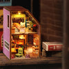 A wooden miniature dollhouse kit featuring a pink and brown structure with an open window displaying small figures and access
