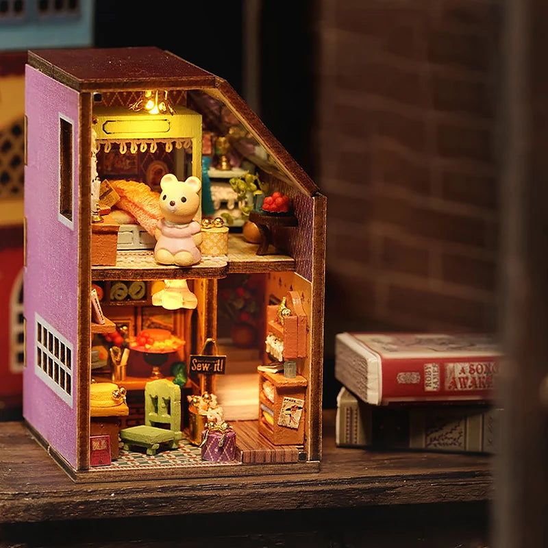 A wooden miniature dollhouse kit featuring a pink and brown structure with an open window displaying small figures and access