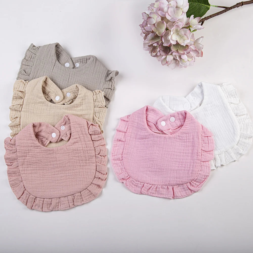 A collection of ruffled muslin cotton baby bibs in shades of beige, pink, and white are displayed on a light background.