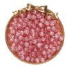 A round dish filled with pink glass crackle beads for jewelry making, set in a gold-toned frame.