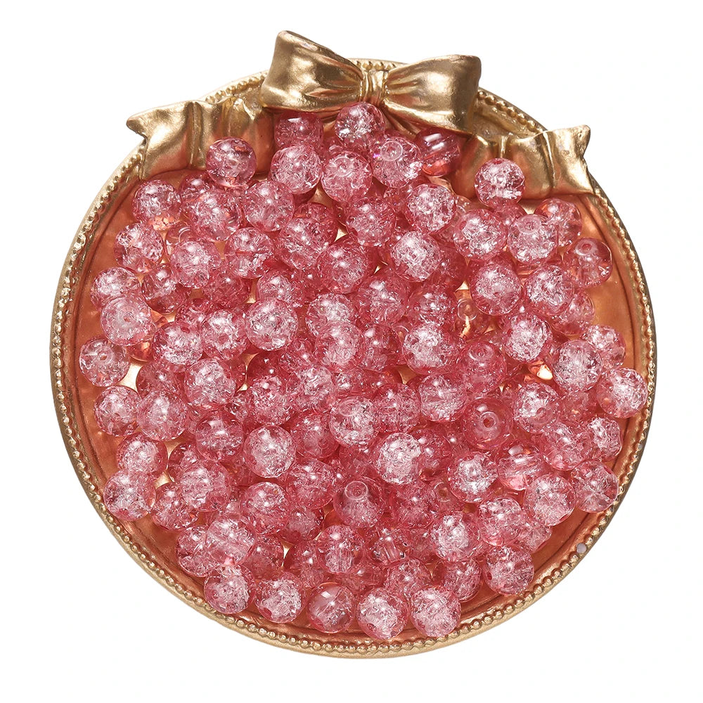 A round dish filled with pink glass crackle beads for jewelry making, set in a gold-toned frame.