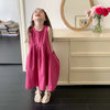 A young girl in a pink sleeveless cotton sundress stands smiling in front of a light wood cabinet and a woven mirror.