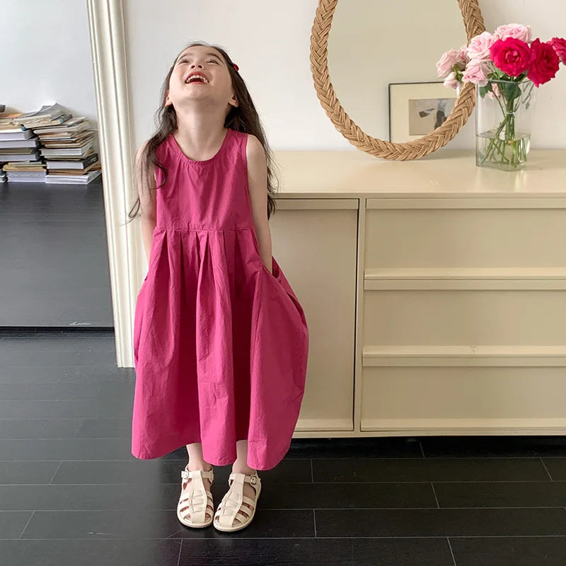 A young girl in a pink sleeveless cotton sundress stands smiling in front of a light wood cabinet and a woven mirror.