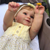A close-up of a lifelike vinyl baby doll with 3D skin detail, wearing a yellow top and a flower headband.