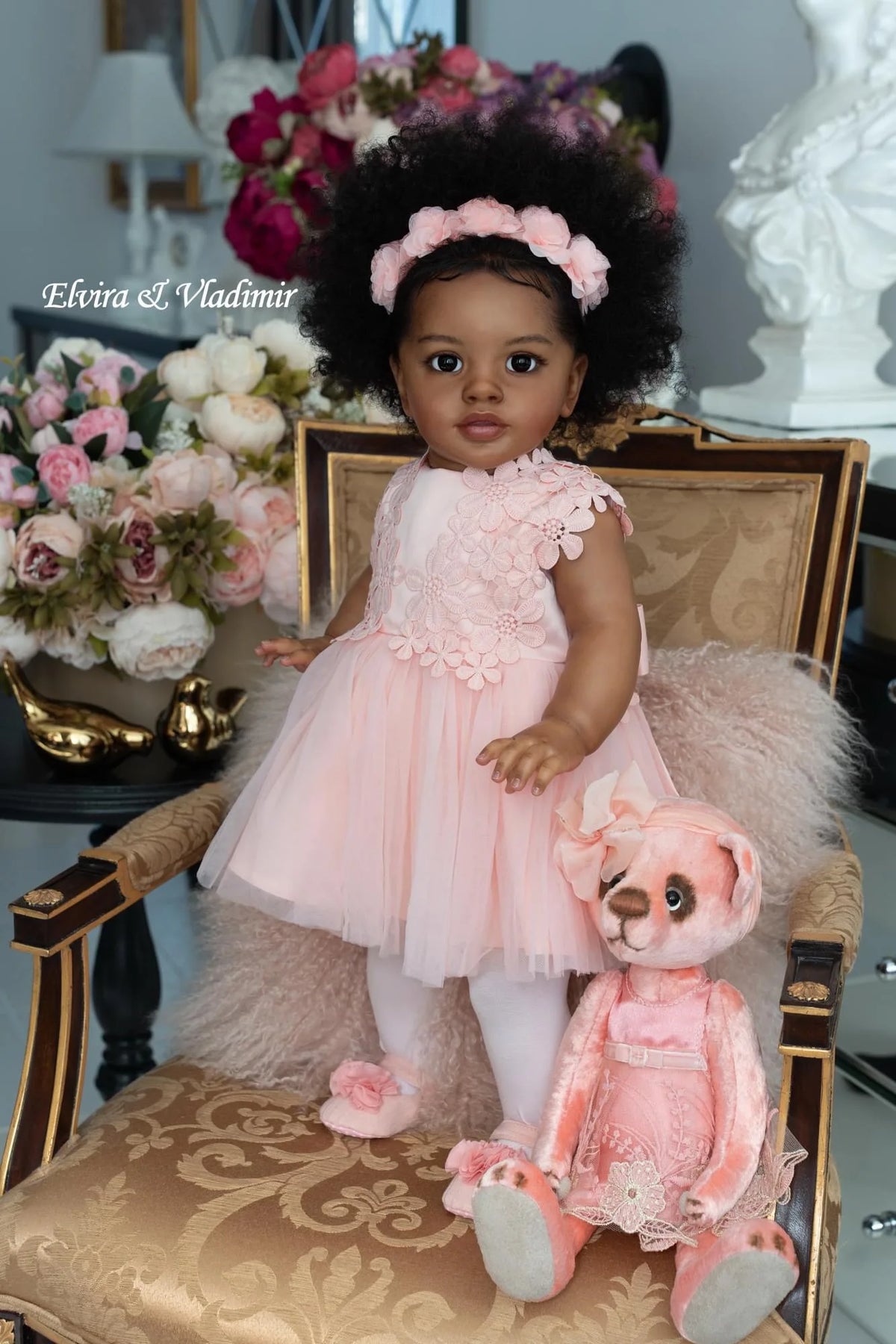 A toddler doll with hand-rooted black hair wearing a pink dress sitting on a chair.
