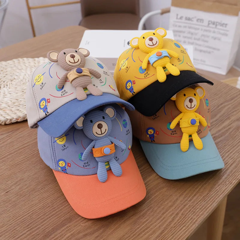 A yellow toddler bear baseball cap with an adjustable strap and a smaller bear decoration resting on a wooden table.