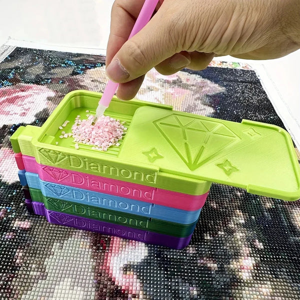 A hand is using a pink tool to apply glitter onto a diamond painting inside a stack of colorful plastic storage trays.