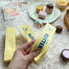 A hand holds a yellow butter stick squishy toy next to various pastel-colored butter products and candies.