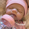 A close-up of a newborn silicone reborn baby doll wearing a pink knitted hat and being fed a pink bottle.