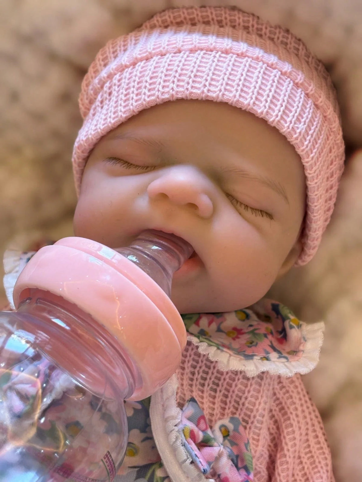 A close-up of a newborn silicone reborn baby doll wearing a pink knitted hat and being fed a pink bottle.