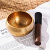 A handcrafted Tibetan singing bowl with a gold finish, silk cushion, and wooden mallet resting on white paper.