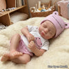 A realistic reborn baby doll with pink knitted hat and clothing lying on a fluffy white surface.