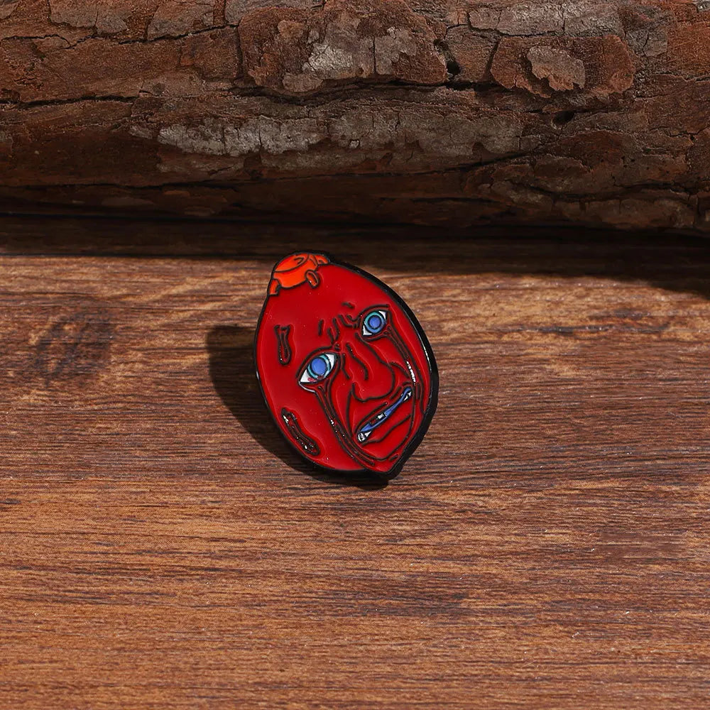 An enamel lapel pin depicting a stylized, distressed face with intense eyes and open mouth on a wooden surface.