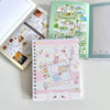 A pink and white spiral notebook filled with cute stickers and illustrations, next to an open green book.