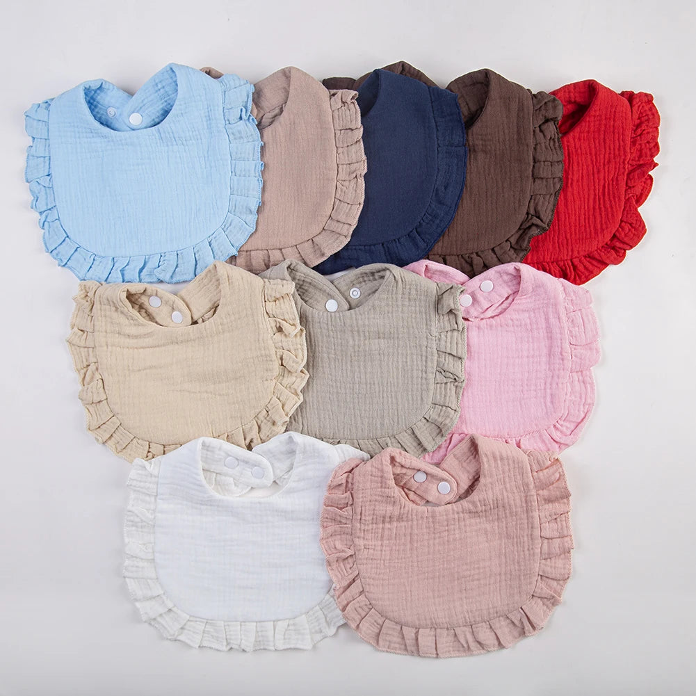 A collection of ruffled muslin cotton baby bibs in various pastel and earthy tones, including blue, beige, gray, pink, and re