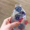 A hand holds a clear glass bottle filled with purple and blue squishy fruit toys.