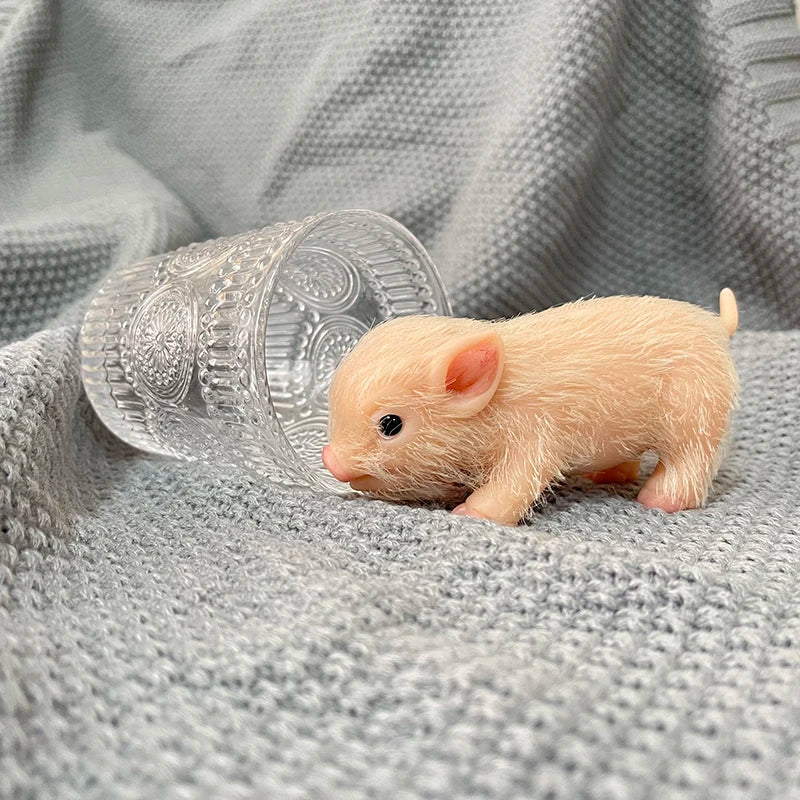 A small, light orange silicone reborn piglet doll lying on a gray fabric next to a clear glass.