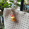 A handmade crochet yellow animal keychain with a red heart charm attached to a light beige woven fabric bag.