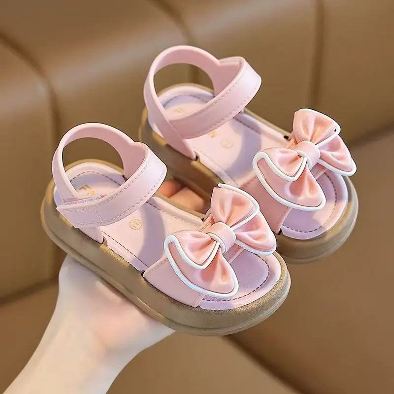 Pair of pink girls summer sandals with bow details shown being held by a hand.